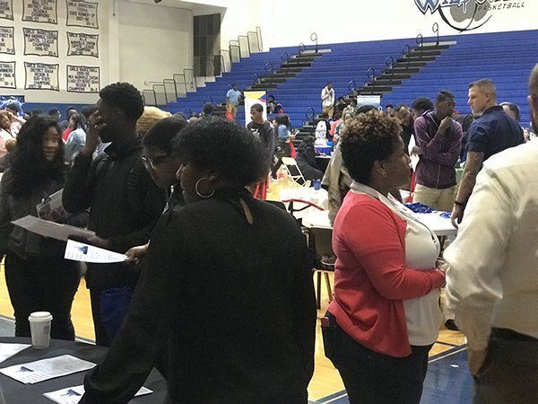 People at tables in a gymnasium, possibly a career fair. Blue bleachers in the background.