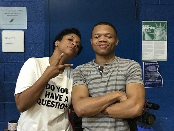 Two people pose for a photo in front of a blue wall. One person makes a peace sign, the other crosses their arms.