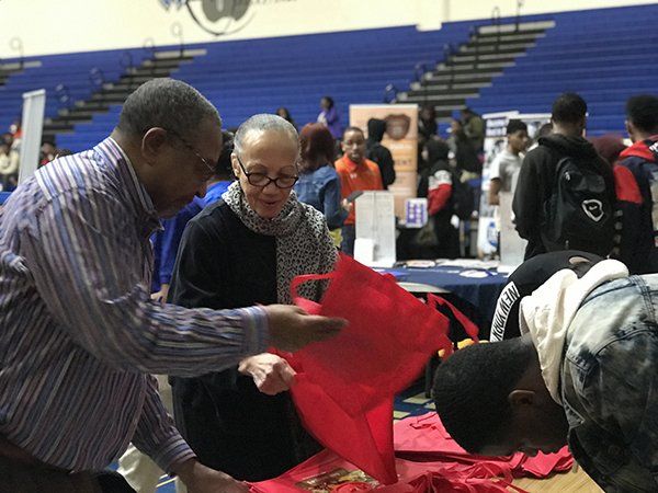 Two people examine a red bag and items at an event table, with other people and tables in the background.