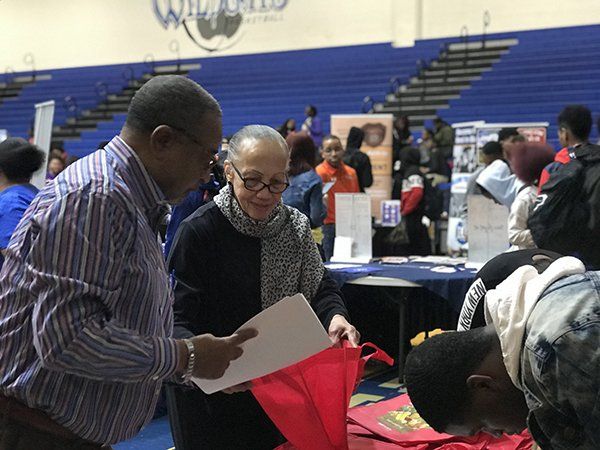 Two people looking at a paper in a red bag at an event, with tables and people in the background.