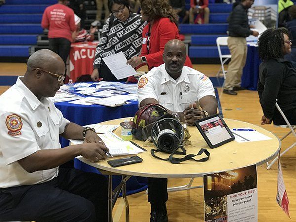 Two firefighters at a table with equipment, answering questions at an event in a gymnasium.