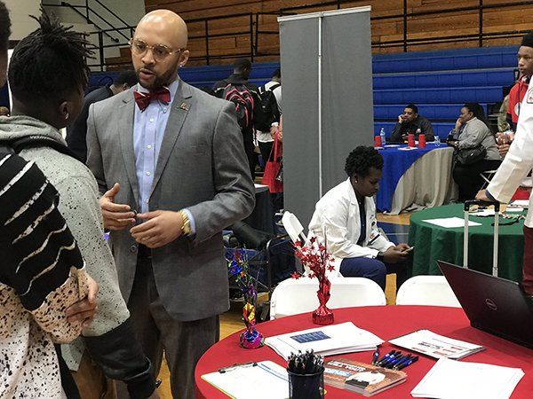 Man in a gray suit speaks to a person at a career fair, red table with documents, people in background.