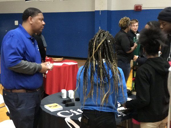 Man in blue shirt talks to people at a table in a gym. Attendees are looking at items on the table.