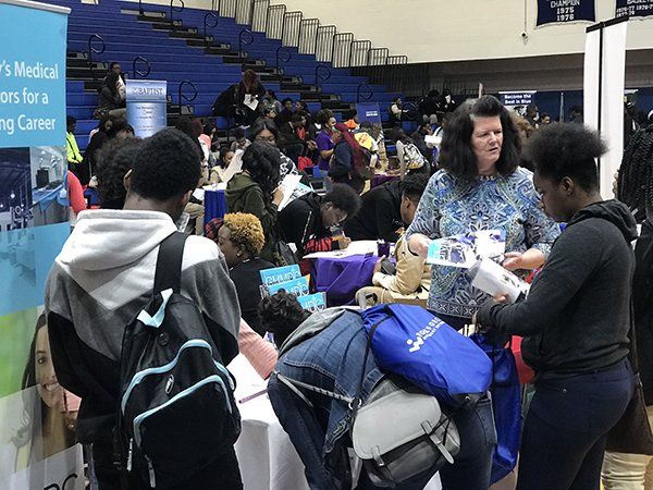 Students at tables at a career fair in a gymnasium setting, looking at materials, and talking to professionals.
