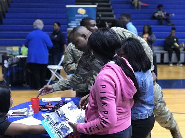People looking at brochures at a table in a gymnasium. Man in uniform, woman in pink hoodie.