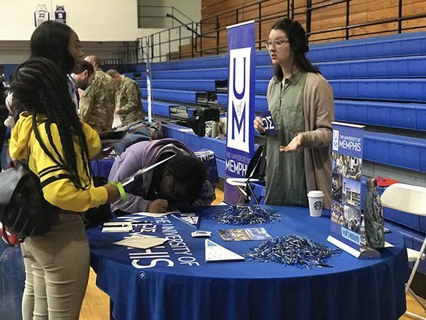 University of Memphis table at an event. A person speaks to two others. Blue table cloth with the university logo.