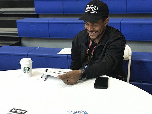 Man smiles while holding papers at a table with pens, a Starbucks cup, and a phone. Blue bleachers in the background.