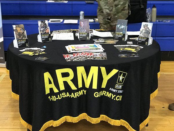 Army recruitment table with brochures and materials, black tablecloth with yellow fringe and 