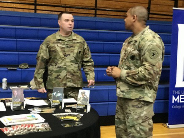 Two soldiers in uniform talking at a table with promotional materials in a gymnasium.