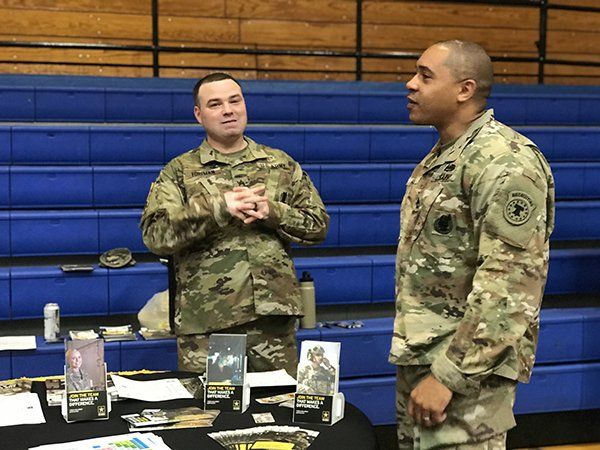 Two men in military uniforms talking at a table in a gymnasium. One is gesturing. Blue bleachers in background.