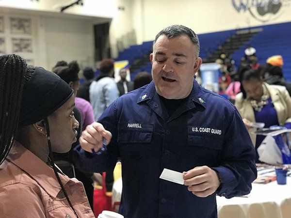 Man in U.S. Coast Guard uniform talks to a person at a recruitment event. They are near a table with materials.