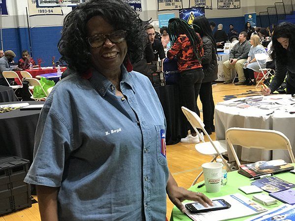 Woman at a table in a crowded event hall, smiling, wearing a blue shirt, looking at a phone.