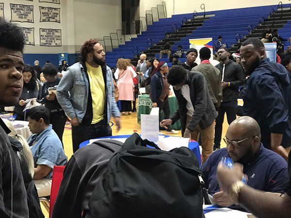 People at a college fair, various booths with brochures and people talking, in a gymnasium with blue seating.