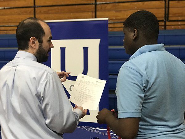 Man in a button-down shirt points at a paper, talking to a person in a light blue polo shirt near a blue banner.
