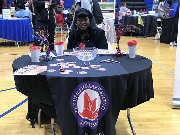 Woman at The Healthcare Institute table, smiling, with promotional items and logo on display. Indoor event setting.
