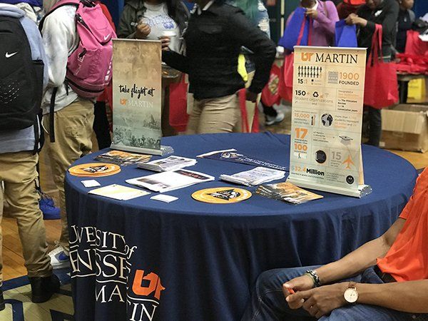 A table with UT Martin information at an event. People in the background, a blue tablecloth, and banners.