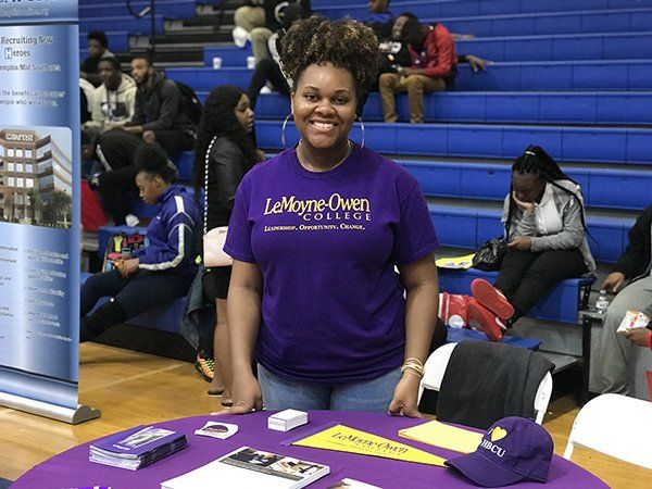 Woman at a college information table wearing a purple shirt, smiling. Event in a gym setting.