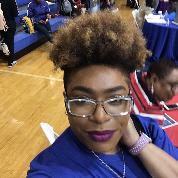 Woman with glasses and short, light-brown curly hair smiles; wears blue shirt, purple lipstick, and a pink watch; indoors.