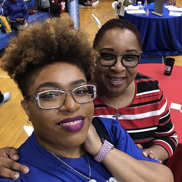 Two women smiling close up. One with curly hair, glasses, and a purple lip. The other in glasses and a striped shirt.