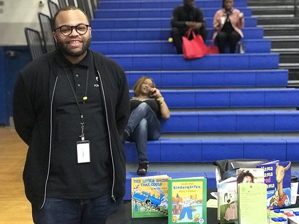 Man with glasses stands by a table of books in a school gym, with people on bleachers in the background.