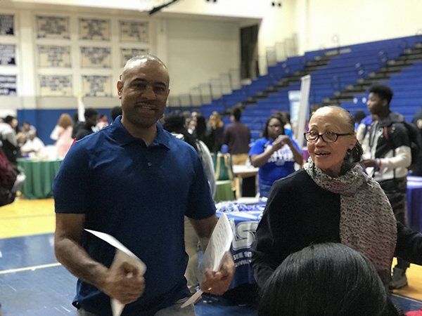 Man and woman smiling at camera in a gymnasium with tables set up for an event; people in the background.