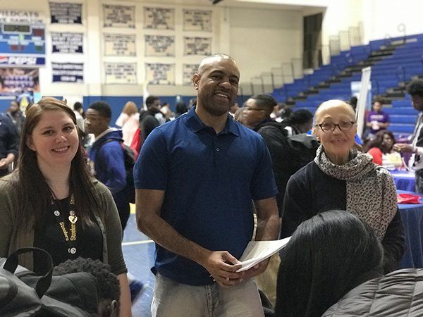 Three adults smiling, standing in a gymnasium with tables and people. The man holds papers.