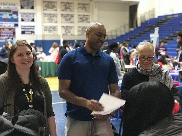 People at event, man holding papers, smiling woman and person looking down. Indoors, blue and green.