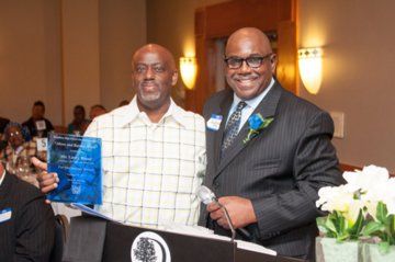 Two men at an event; one holds a blue award, other wears a suit. Event setting with a podium.