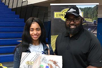 Woman and man posing next to a banner. Woman holds a painting. Blue bleachers are in the background.