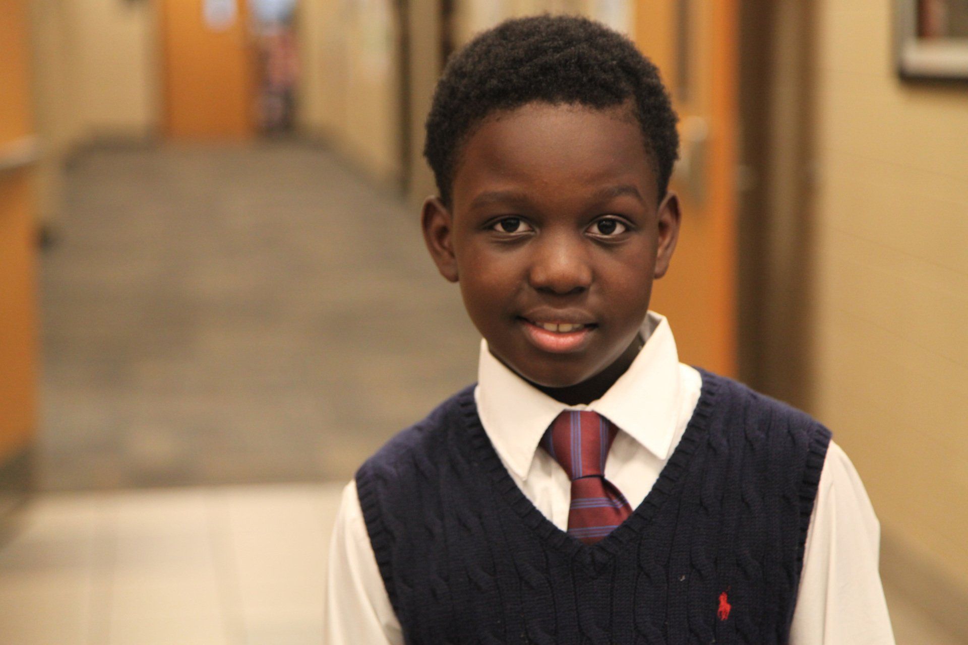 Young Black boy smiles in school uniform in a hallway.