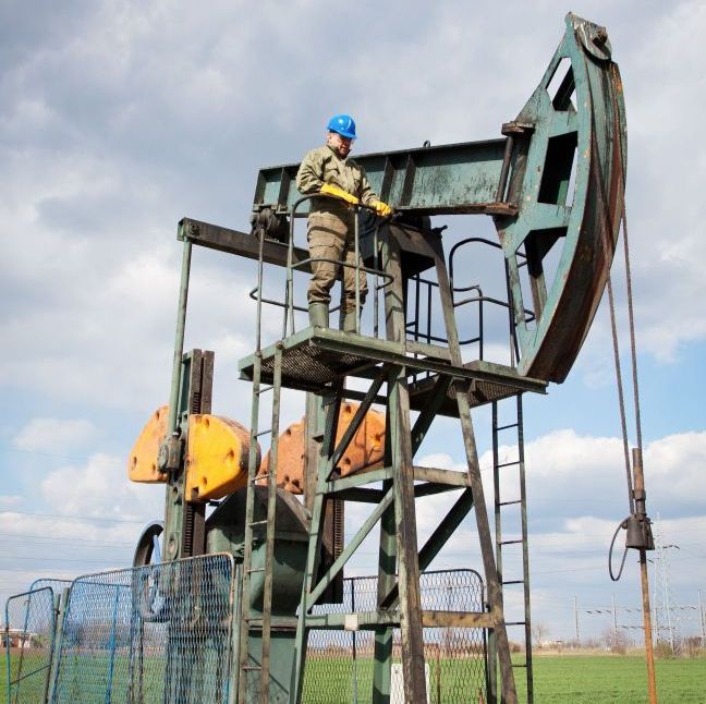 Worker on oil pump platform, wearing blue hard hat and gloves, in a field under a partly cloudy sky.