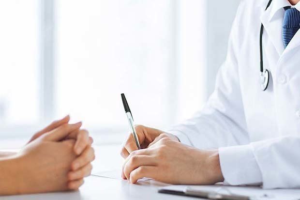Un medico in camice bianco scrive su un documento mentre è seduto a una scrivania con un paziente durante una visita medica.