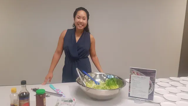 Woman standing behind a table with a salad bowl and recipe card, smiling.