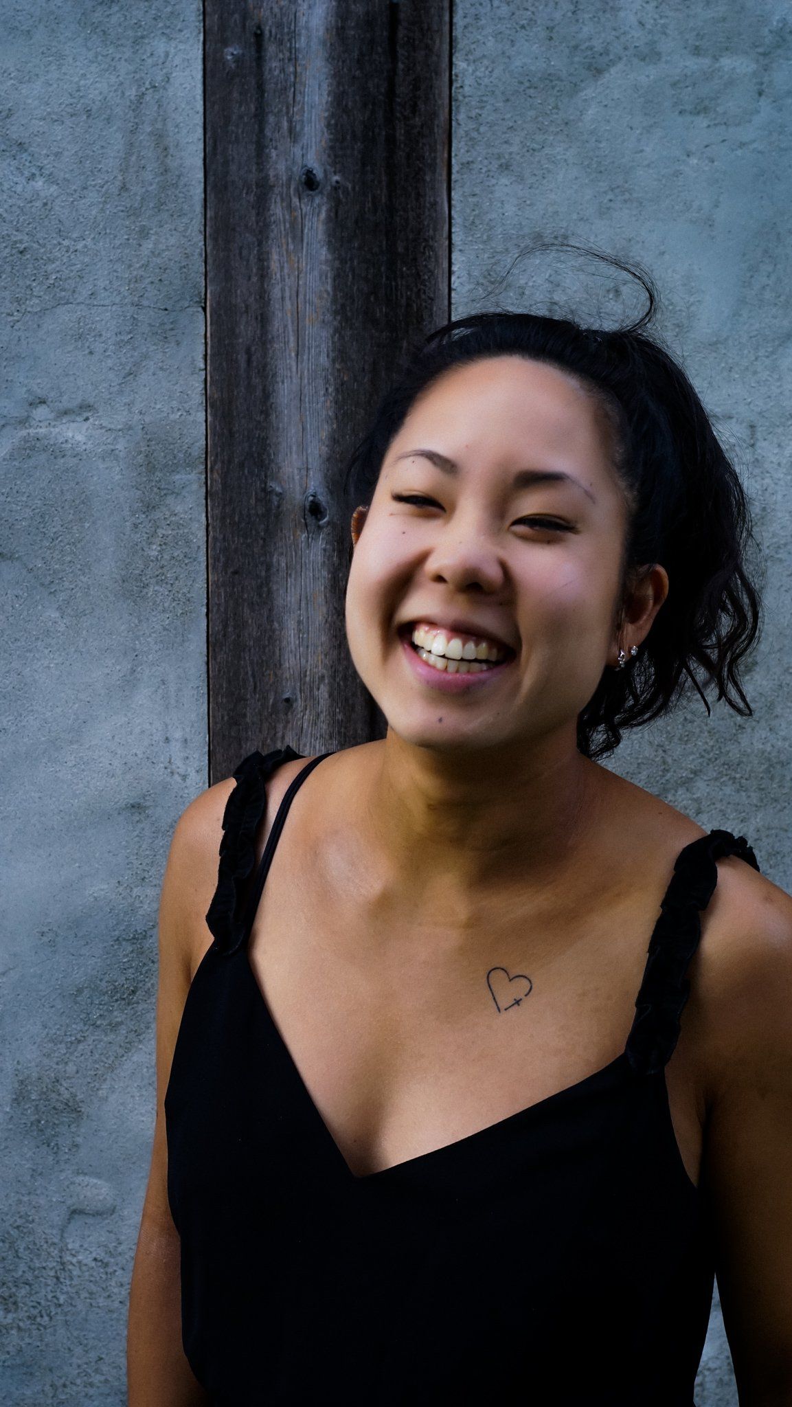 Smiling person in a black tank top, heart tattoo on her chest, against a gray wall with wooden beam.