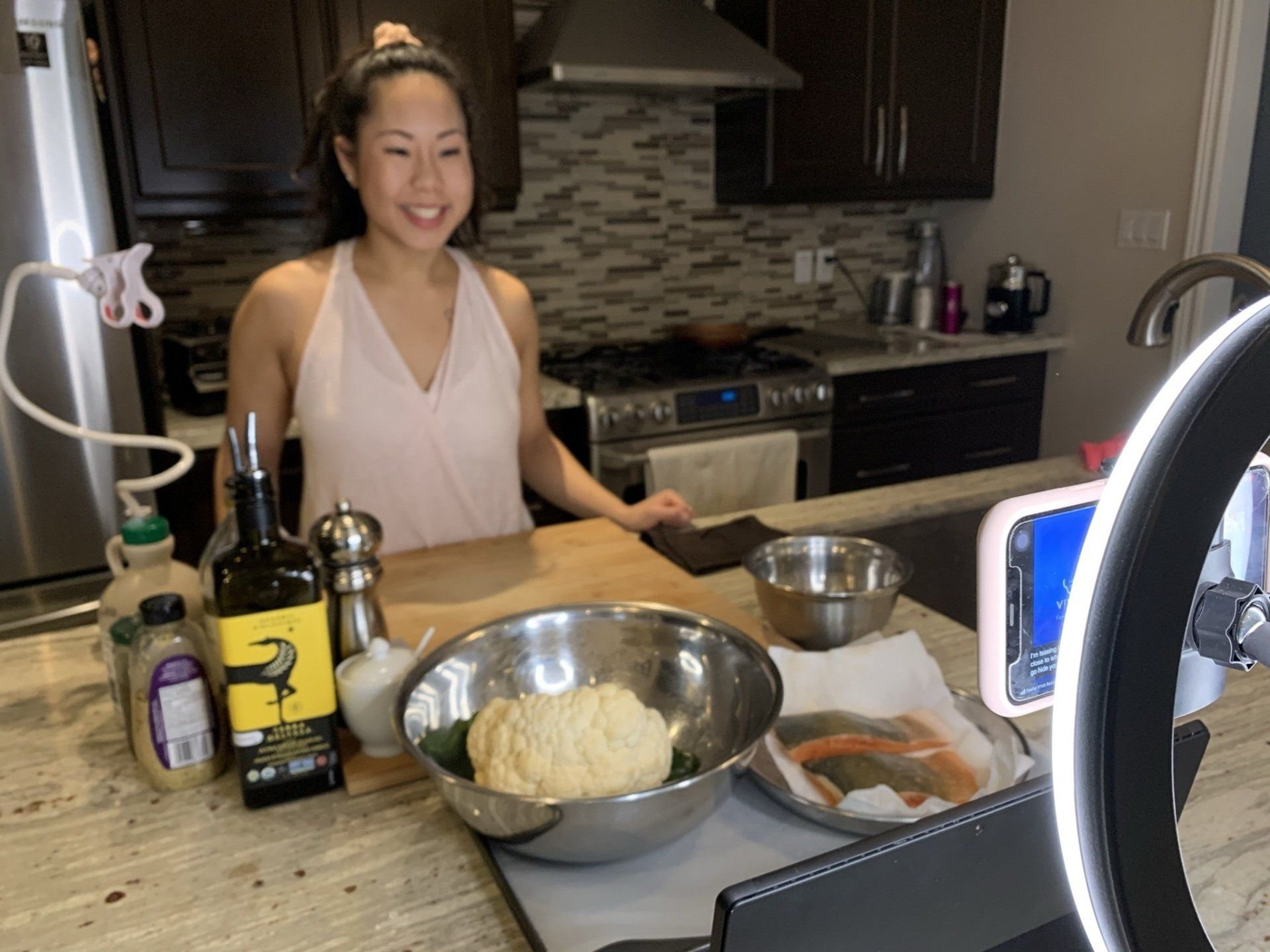 Woman cooking in kitchen, smiling, with ingredients and phone setup for recording.