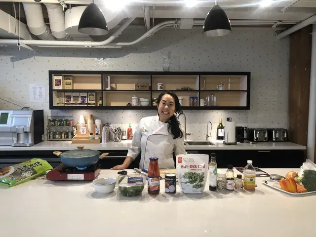 Chef smiles in a modern kitchen, ingredients laid out. A pot simmers on a portable stove.