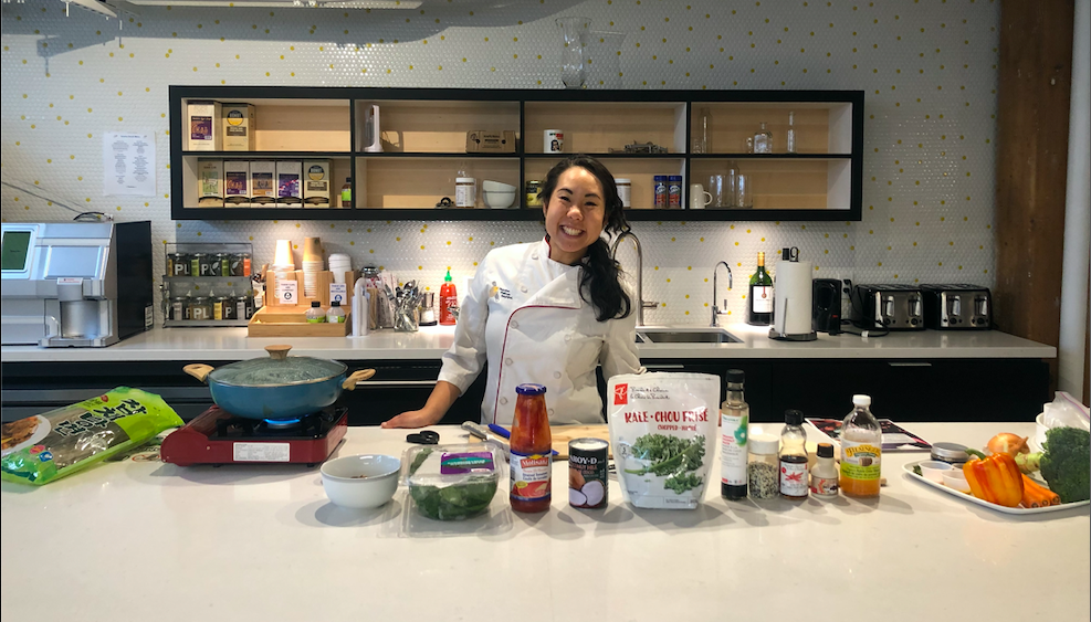 Woman in chef's coat smiles in a kitchen, ingredients and a pot on the counter.