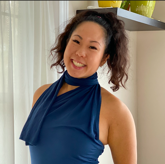 Woman with dark hair smiles, wearing a navy blue halter top, standing in front of a window with white curtains.