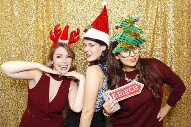 Three women in festive headwear pose in front of a gold sequin backdrop; smiling, holding a 