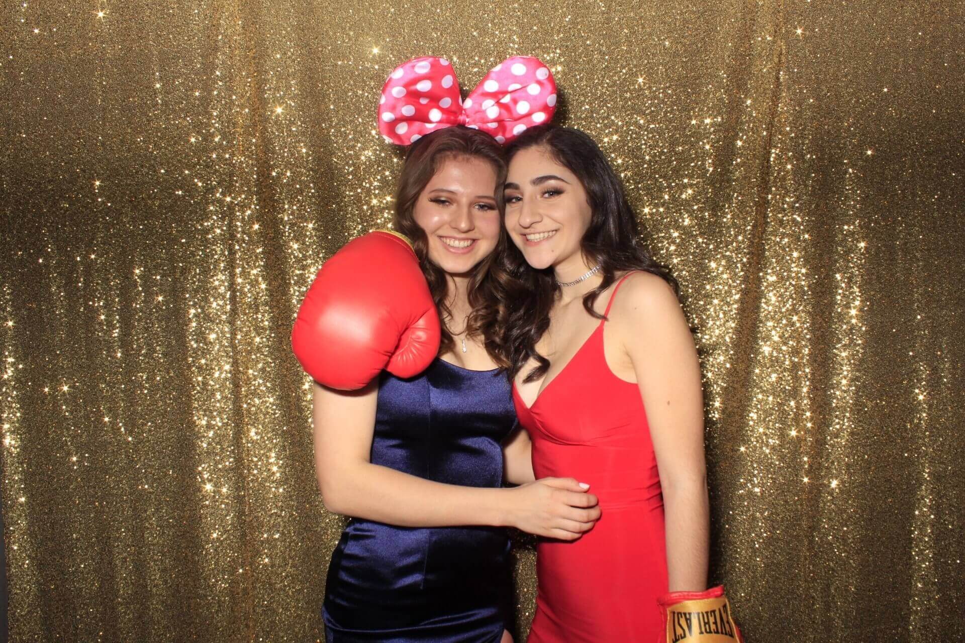 Two people at a photo booth with props: boxing gloves and a large bow. Gold sequin backdrop.