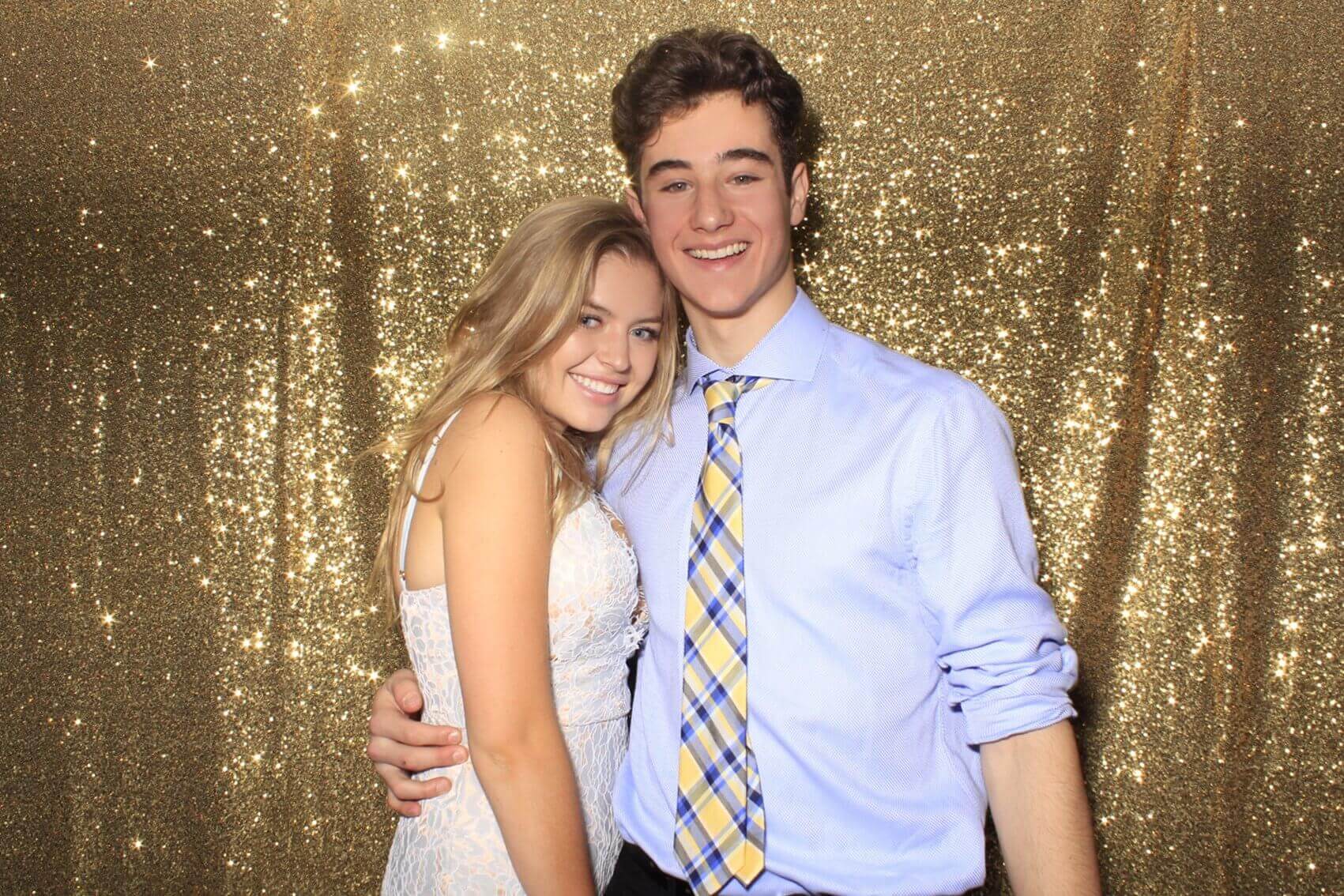 Couple poses in front of a sparkly gold backdrop. Smiling, woman in white dress, man in blue shirt and tie.