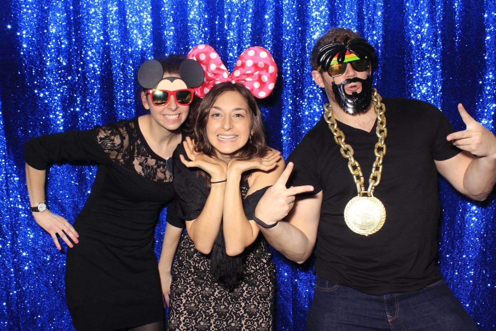 Three people posing with photo booth props in front of a blue sequin backdrop; wearing sunglasses, mouse ears, and gold chain.