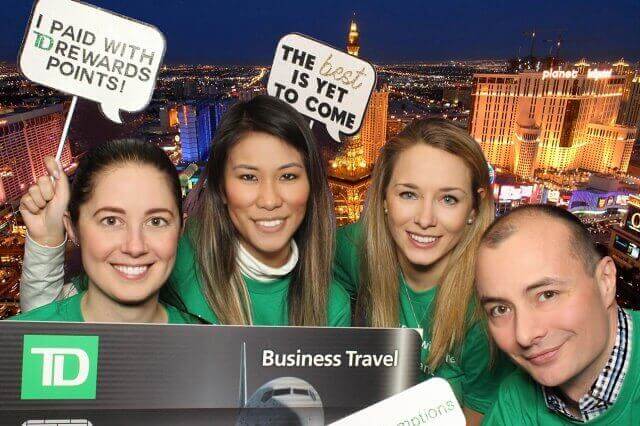 Four people in green shirts pose with props and a Las Vegas backdrop for a TD Business Travel promotion.