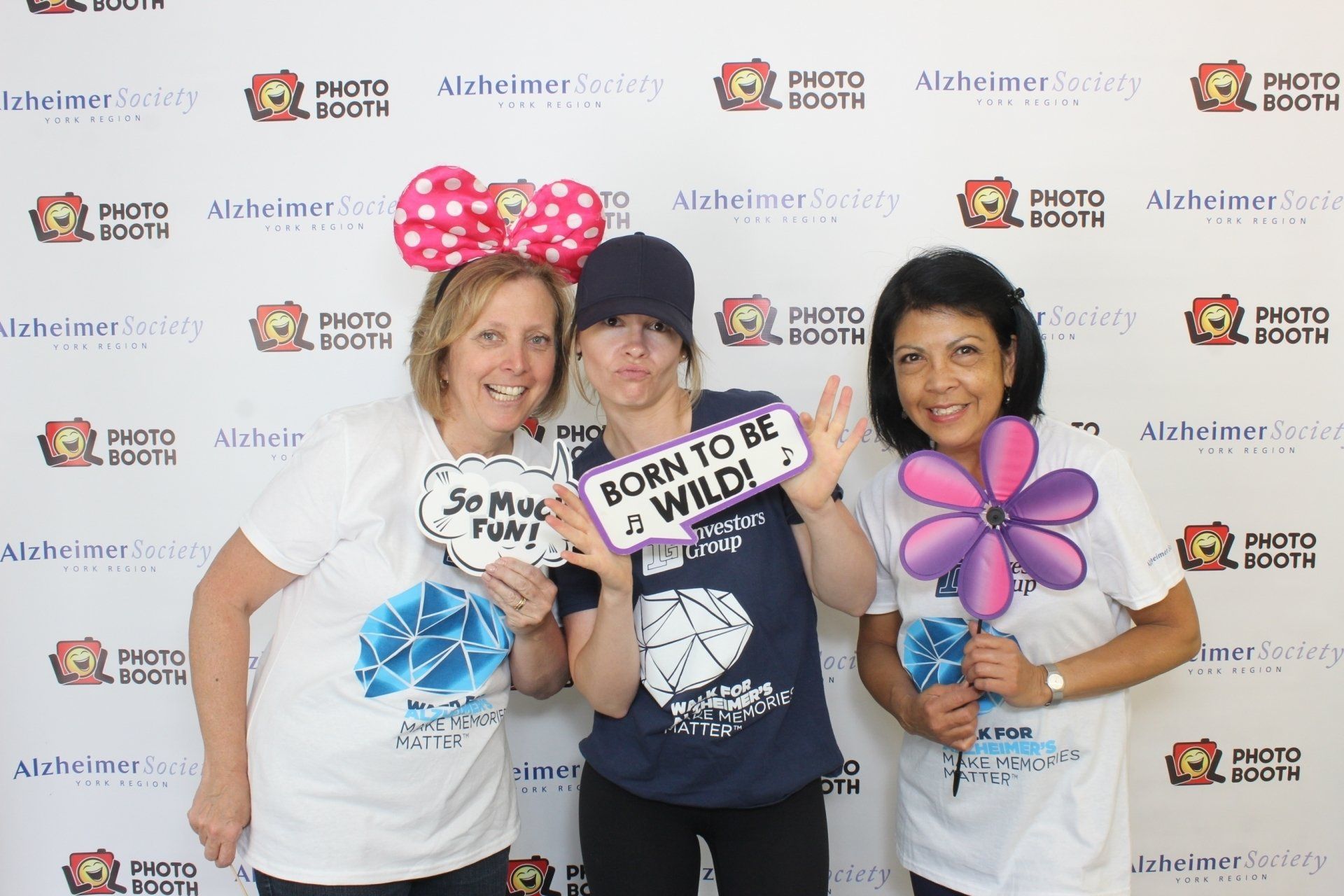 Three women pose at an Alzheimer's event photo booth, holding props. Background shows event branding.