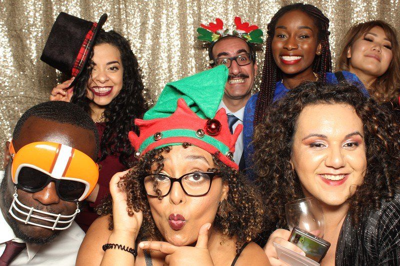 Group of people in a photo booth, smiling and wearing festive props against a gold backdrop.