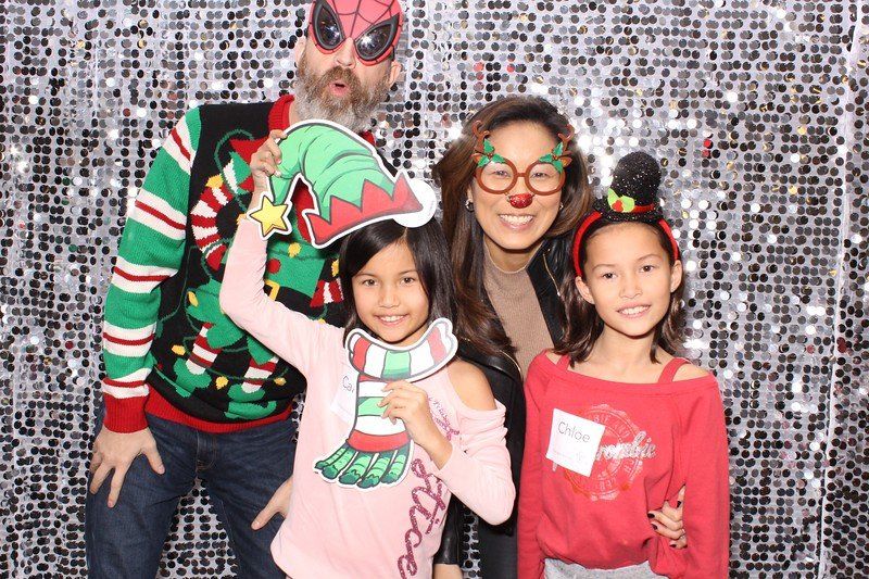 Mom, dad and two daughters posing in a photo booth with a silver background with holiday props.