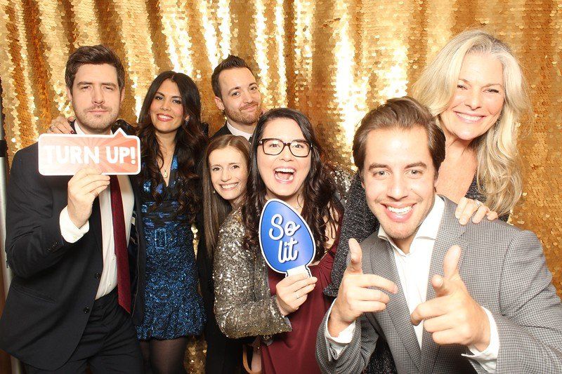 Group photo at a party with a gold sequin backdrop. People are smiling, holding props, and pointing.