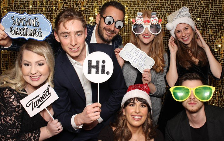 Group of eight people posing in a photo booth; holding props like signs and funny glasses in front of a gold sequined backdrop.