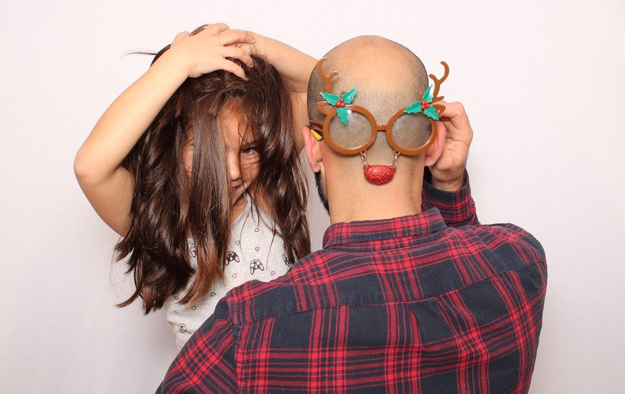 Man wearing reindeer glasses, child behind him, both in front of a white backdrop.