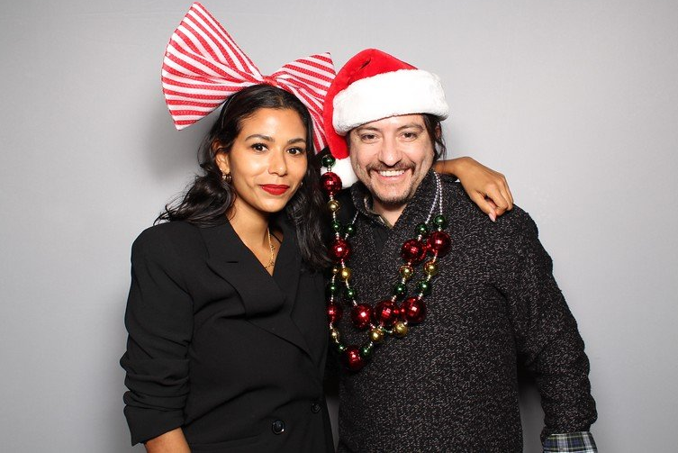 Woman in black blazer and large striped bow headband, man in Santa hat and sweater, arm around woman.
